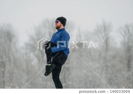 Male athlete in blue jacket and black leggings performs a stretching exercise outdoors in a snowy landscape with trees and falling snowflakes in the background 140033009