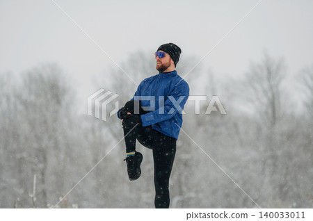 Male athlete in blue jacket and black leggings performs warm-up exercise outdoors in snowy winter landscape with trees and soft snowfall in the background Male athlete in blue jacket and black leggings performs warm-up exercise outdoors in snowy winter landscape with trees and soft snowfall in the background 140033011
