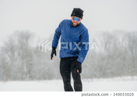 Male athlete wearing blue jacket and sunglasses runs through snowy landscape during winter, snowflakes falling and trees blurred in the background Male athlete wearing blue jacket and sunglasses runs through snowy landscape during winter, snowflakes falling and trees blurred in the background 140033020