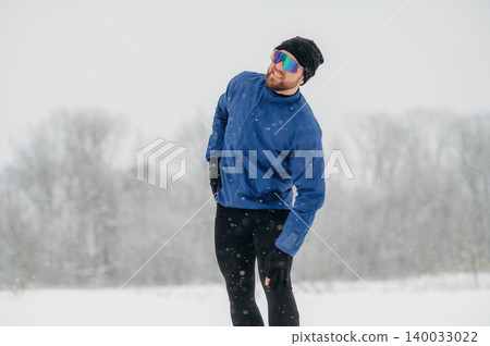 Male athlete wearing blue jacket and sunglasses stretches during winter workout in snowy landscape with trees and soft snowfall in the background Male athlete wearing blue jacket and sunglasses stretches during winter workout in snowy landscape with trees and soft snowfall in the background 140033022