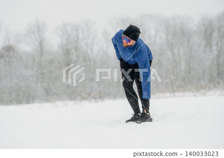 Male runner in blue jacket and black leggings stretches while jogging in snowy landscape with trees and falling snowflakes in the background 140033023