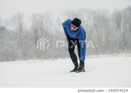 Male runner in blue jacket and black leggings stretches while kneeling on snow-covered ground during a snowstorm in a winter landscape with trees in the background Male runner in blue jacket and black leggings stretches while kneeling on snow-covered ground during a snowstorm in a winter landscape with trees in the background 140033024