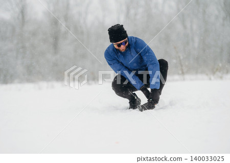 Male hiker in blue jacket and black pants crouches in deep snow, adjusting gear while surrounded by a snowy landscape and trees in the background Male hiker in blue jacket and black pants crouches in deep snow, adjusting gear while surrounded by a snowy landscape and trees in the background 140033025