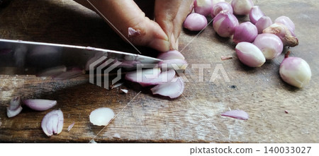 The process of cutting fresh shallots using a knife on a wooden chopping board from a close distance. The process of cutting fresh shallots using a knife on a wooden chopping board from a close distance. 140033027