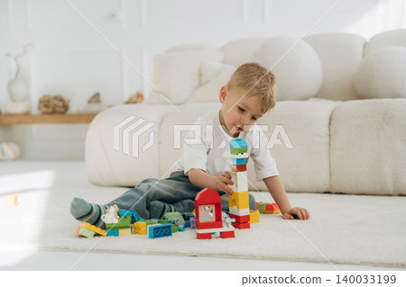 Young boy playing with colorful building blocks on a soft rug in a bright, modern living room with a cozy sofa and decorative items in the background Young boy playing with colorful building blocks on a soft rug in a bright, modern living room with a cozy sofa and decorative items in the background 140033199