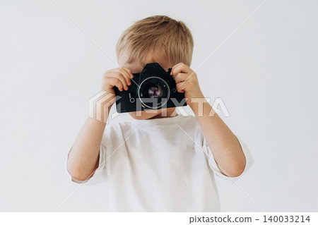 Young child holding a camera in front of their face, wearing a white t-shirt, with a plain light background emphasizing the subject's playful engagement with photography Young child holding a camera in front of their face, wearing a white t-shirt, with a plain light background emphasizing the subject's playful engagement with photography 140033214