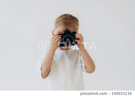 Young boy holding a digital camera in front of his face, wearing a white t-shirt, with a plain light background creating a clean and simple composition Young boy holding a digital camera in front of his face, wearing a white t-shirt, with a plain light background creating a clean and simple composition 140033216