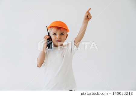 Young boy wearing an orange cap holds a walkie-talkie while pointing upwards, dressed in a white t-shirt against a plain white background Young boy wearing an orange cap holds a walkie-talkie while pointing upwards, dressed in a white t-shirt against a plain white background 140033218