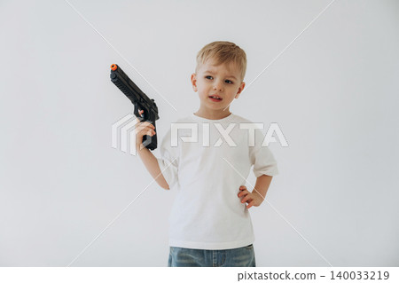 Young boy holding a toy gun in a white t-shirt and denim jeans, standing confidently against a plain white background Young boy holding a toy gun in a white t-shirt and denim jeans, standing confidently against a plain white background 140033219