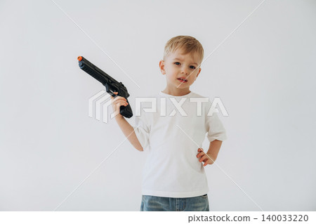 Young boy in white t-shirt and blue jeans holds a toy gun with an orange tip, posing against a plain white background in a playful manner Young boy in white t-shirt and blue jeans holds a toy gun with an orange tip, posing against a plain white background in a playful manner 140033220