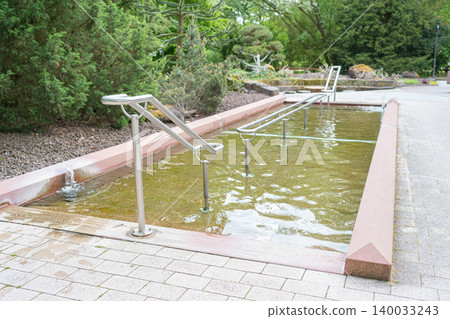 An outdoor Kneipp water cure therapy pool for feet walking wellness treatments located in a beautiful green spa park. Concrete construction filled with cold fresh water and fitted with metal railings An outdoor Kneipp water cure therapy pool for feet walking wellness treatments located in a beautiful green spa park. Concrete construction filled with cold fresh water and fitted with metal railings 140033243