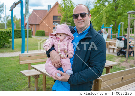 A happy loving father wearing sunglasses holds his small child dressed in a pink warm jumpsuit on an outdoor public playground. Concept of happy parenthood, dad life, and family weekend 140033250