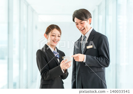 A businessman and woman in suits talking while looking at their smartphones (communication) A businessman and woman in suits talking while looking at their smartphones (communication) 140033541