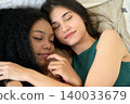Two young women resting together while holding hands in tender moment of friendship and connection while lying on bed with eyes closed 140033679