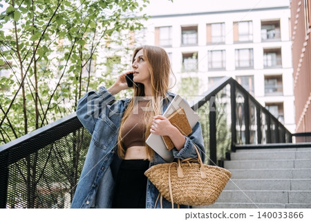 Young woman talks on smartphone while walking down outdoor city stairs holding laptop. Agile workflow execution, fast paced business, independent contractor workflow. Young woman talks on smartphone while walking down outdoor city stairs holding laptop. Agile workflow execution, fast paced business, independent contractor workflow. 140033866