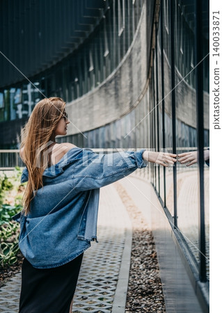 Woman standing outdoors next to glass building touching her reflection. Connecting with architecture, urban mindfulness concept, finding peace in city. 140033871