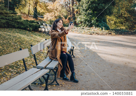 Woman sits on bench in public park while taking selfie or recording video with smartphone. User generated content UGC production, mobile photography in nature, social media marketing imagery. Woman sits on bench in public park while taking selfie or recording video with smartphone. User generated content UGC production, mobile photography in nature, social media marketing imagery. 140033881