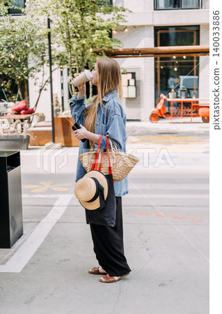 Young woman drinks from cup while holding smartphone on urban street. Zero waste daily routine, sustainable textile accessories, conscious retail choices. Young woman drinks from cup while holding smartphone on urban street. Zero waste daily routine, sustainable textile accessories, conscious retail choices. 140033886
