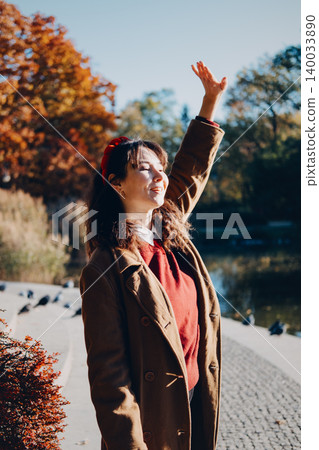 Woman stands smiling with raised hand in autumn park by pond. Importance of vitamin D autumn and winter, high risk of vitamin D deficiency for woman. Woman stands smiling with raised hand in autumn park by pond. Importance of vitamin D autumn and winter, high risk of vitamin D deficiency for woman. 140033890