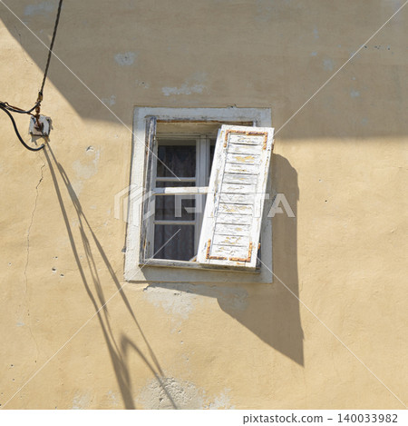 A partially open, weathered white shutter hangs from a window on a pale yellow stucco wall in Piran, Slovenia A partially open, weathered white shutter hangs from a window on a pale yellow stucco wall in Piran, Slovenia 140033982