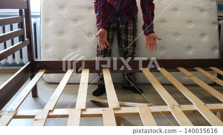 A man stands in a room with his arms outstretched against the backdrop of a raised white mattress in front of a wooden bed with a broken board in the frame, broken furniture, A man stands in a room with his arms outstretched against the backdrop of a raised white mattress in front of a wooden bed with a broken board in the frame, broken furniture, 140035616