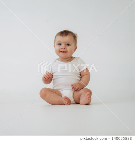 Baby sitting on a light-colored floor wearing a white onesie, smiling and looking directly at the camera with a neutral background Baby sitting on a light-colored floor wearing a white onesie, smiling and looking directly at the camera with a neutral background 140035888