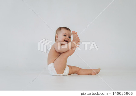 Baby sitting on a white background, playfully holding one foot while wearing a diaper, showcasing a joyful expression and natural curiosity in a minimalistic setting 140035906