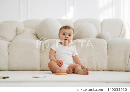 Infant child sitting on a soft rug in a bright room, wearing a white onesie, with a plush sofa and scattered toys visible in the background 140035910