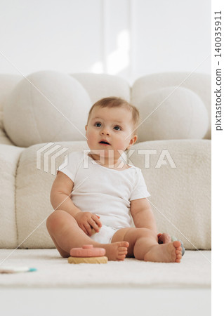 Baby sitting on soft rug in bright room with plush sofa, wearing white onesie, looking curiously at colorful toy in front of him, natural light illuminating the scene 140035911
