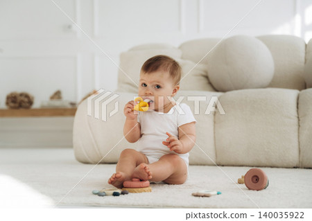 Infant child sitting on soft rug in bright room, playing with colorful toys, plush sofa and wooden decor visible in the background Infant child sitting on soft rug in bright room, playing with colorful toys, plush sofa and wooden decor visible in the background 140035922