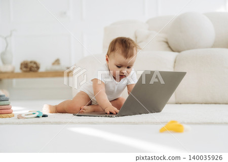 Infant child in white onesie interacts with laptop on soft rug in bright living room, with colorful toys scattered nearby and a cozy sofa in the background Infant child in white onesie interacts with laptop on soft rug in bright living room, with colorful toys scattered nearby and a cozy sofa in the background 140035926