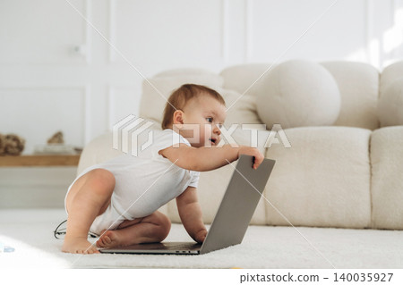 Infant child in white onesie interacts with laptop on soft carpeted floor in bright living room with modern furniture and neutral color palette Infant child in white onesie interacts with laptop on soft carpeted floor in bright living room with modern furniture and neutral color palette 140035927