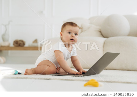 Baby in white onesie sits on soft rug, playing with laptop in bright living room, light-colored sofa and decorative items visible in the background Baby in white onesie sits on soft rug, playing with laptop in bright living room, light-colored sofa and decorative items visible in the background 140035930
