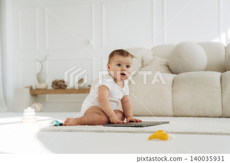 Baby in white onesie sits on soft carpet beside laptop and colorful toys in bright, modern living room with minimalist decor and natural light 140035931