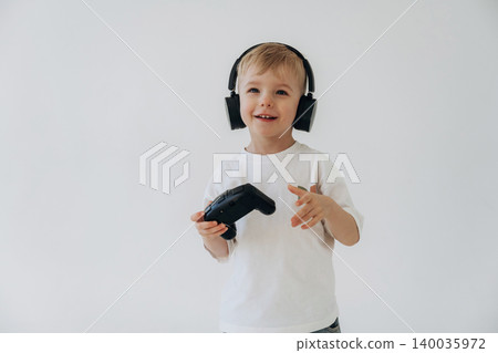 Young boy wearing black headphones holds a video game controller while smiling against a plain white background, showcasing a playful gaming atmosphere Young boy wearing black headphones holds a video game controller while smiling against a plain white background, showcasing a playful gaming atmosphere 140035972