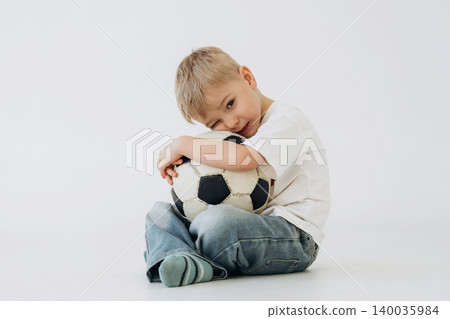 Young boy with blonde hair sitting on the floor, hugging a black and white soccer ball while wearing a white t-shirt and blue jeans against a plain background Young boy with blonde hair sitting on the floor, hugging a black and white soccer ball while wearing a white t-shirt and blue jeans against a plain background 140035984