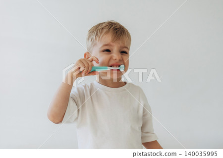 Young boy with light blonde hair brushes his teeth with a blue toothbrush while wearing a white t-shirt against a plain light background Young boy with light blonde hair brushes his teeth with a blue toothbrush while wearing a white t-shirt against a plain light background 140035995