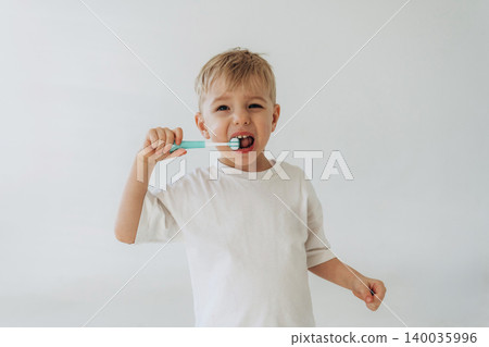 Young boy with light brown hair brushes his teeth with a blue toothbrush, wearing a white t-shirt, standing against a plain light background Young boy with light brown hair brushes his teeth with a blue toothbrush, wearing a white t-shirt, standing against a plain light background 140035996