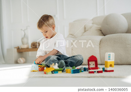 Young boy playing with colorful building blocks on a light carpet in a bright living room, soft sofa and decorative items visible in the background Young boy playing with colorful building blocks on a light carpet in a bright living room, soft sofa and decorative items visible in the background 140036005