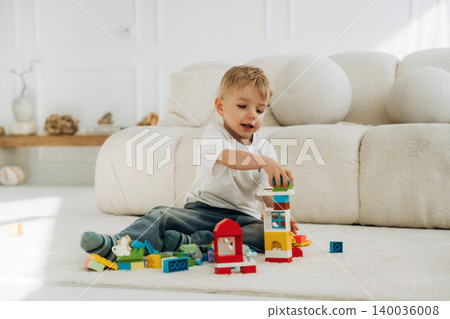 Young boy with light brown hair stacking colorful building blocks on a soft rug in a bright living room with a cozy sofa and natural light Young boy with light brown hair stacking colorful building blocks on a soft rug in a bright living room with a cozy sofa and natural light 140036008