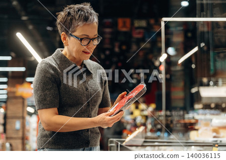 Middle-aged woman with short hair wearing glasses examines a book in a modern bookstore, shelves filled with various items visible in the background 140036115