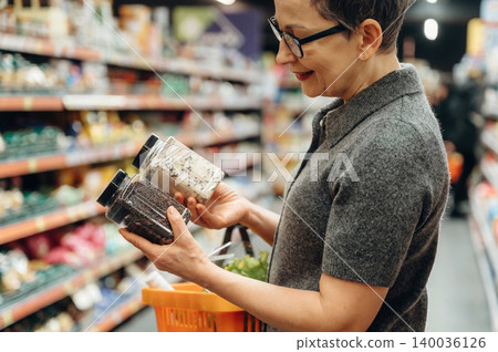 Middle-aged woman with short hair wearing glasses compares two jars of spices while shopping in a grocery store aisle filled with various products and shelves Middle-aged woman with short hair wearing glasses compares two jars of spices while shopping in a grocery store aisle filled with various products and shelves 140036126