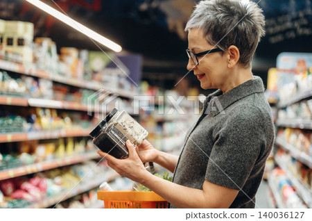 Mature woman with short gray hair examines spice jars while shopping in a grocery store aisle, colorful products displayed on shelves in the background Mature woman with short gray hair examines spice jars while shopping in a grocery store aisle, colorful products displayed on shelves in the background 140036127