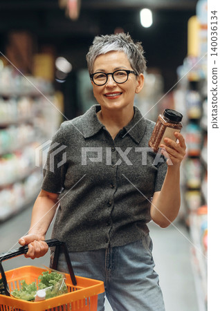 Senior woman with short gray hair wearing glasses holds a spice jar and shopping basket while standing in a grocery store aisle filled with products Senior woman with short gray hair wearing glasses holds a spice jar and shopping basket while standing in a grocery store aisle filled with products 140036134