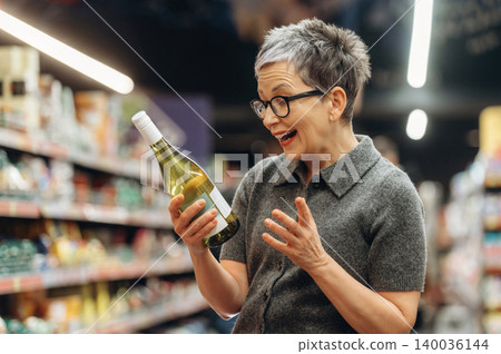 Middle-aged woman with short gray hair and glasses holds a bottle of white wine, smiling in a grocery store aisle filled with various products and shelves Middle-aged woman with short gray hair and glasses holds a bottle of white wine, smiling in a grocery store aisle filled with various products and shelves 140036144