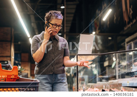 Elderly Asian woman talking on smartphone while shopping at a grocery store, inspecting fresh meat displayed in a glass case with shopping cart nearby 140036181