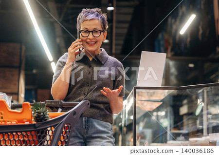 Senior woman with short gray hair talking on smartphone while standing in grocery store aisle, shopping cart filled with groceries, bright lighting and shelves in background Senior woman with short gray hair talking on smartphone while standing in grocery store aisle, shopping cart filled with groceries, bright lighting and shelves in background 140036186