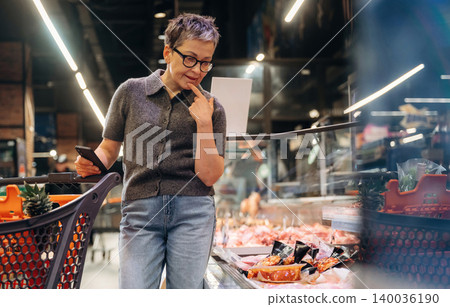 Middle-aged woman with short gray hair using smartphone while shopping for groceries in a modern supermarket with colorful displays of fresh produce and packaged items Middle-aged woman with short gray hair using smartphone while shopping for groceries in a modern supermarket with colorful displays of fresh produce and packaged items 140036190