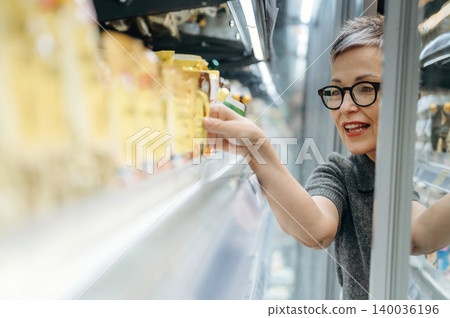 Middle-aged woman with short gray hair wearing glasses selects packaged food items from a refrigerated display in a grocery store aisle with bright lighting Middle-aged woman with short gray hair wearing glasses selects packaged food items from a refrigerated display in a grocery store aisle with bright lighting 140036196