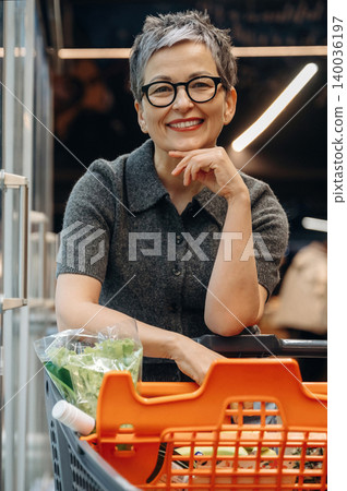 Mature Asian woman with short gray hair and glasses smiles while leaning on a shopping cart filled with fresh vegetables in a modern grocery store setting Mature Asian woman with short gray hair and glasses smiles while leaning on a shopping cart filled with fresh vegetables in a modern grocery store setting 140036197
